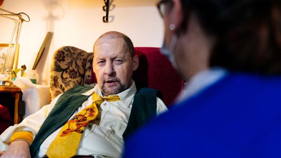 Patient sitting in an arm chair at home being visited by a NHS nurse.