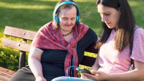 Two women on a park bench, one wearing headphones while the other holds the device.