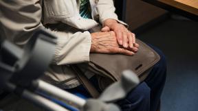 A seated elderly patient resting her hands on a handbag.