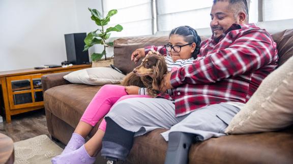 A parent wearing prosthetic legs, sitting on the sofa next to his daughter and their dog.