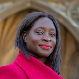 A photo of Abena Oppong-Asare, wearing a red coat, standing in front of a blurred image of what may be the Houses of Parliament in London.