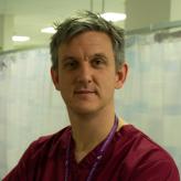 Accessibility description: A photo of Dr Chris Pickering, a white middle aged man with short spiky hair, clean shaven, wearing dark red surgical scrubs, an NHS lanyard and turning to face the camera.