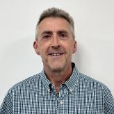 A white middle-aged man with short grey hair wears a grey and white check shirt and stands smiling in front of a white background