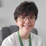 Accessibility description: A photo of Zoe Mitchell, a young white woman with short dark brown hair, seated on a chair, wearing glasses and smiling towards the camera.