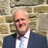 Accessibility description: A photo of Dr Mark Buchanan, a white man with short gray hair and a beard, smiling towards the camera, wearing a suit.